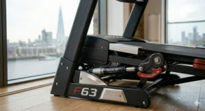 A photorealistic, high-detail 4K photograph capturing the detailed hydraulic folding mechanism, frame details, and space-saving features of the Sole F63 treadmill in its partially folded state within a London apartment home gym.