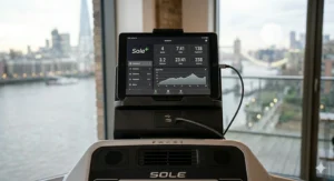 A photorealistic, high-detail 4K close-up photograph of the Sole F63 treadmill console, focusing on the integrated tablet holder holding an iPad, with the USB charging port and connected cable visible. The background shows the London skyline from the apartment gym.