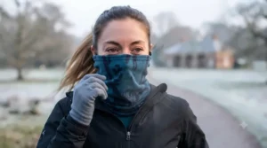 A runner pulling up a windproof neck gaiter over her nose and mouth to protect against biting winter winds on a frosty park path.